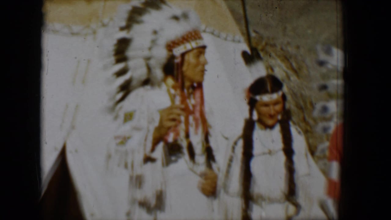 A man in a large feathered headdress stands beside a person with braids near a teepee.