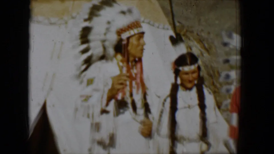 1958 Tucson frame with a man in a large white feathered headdress and another person in white regalia beside a teepee.