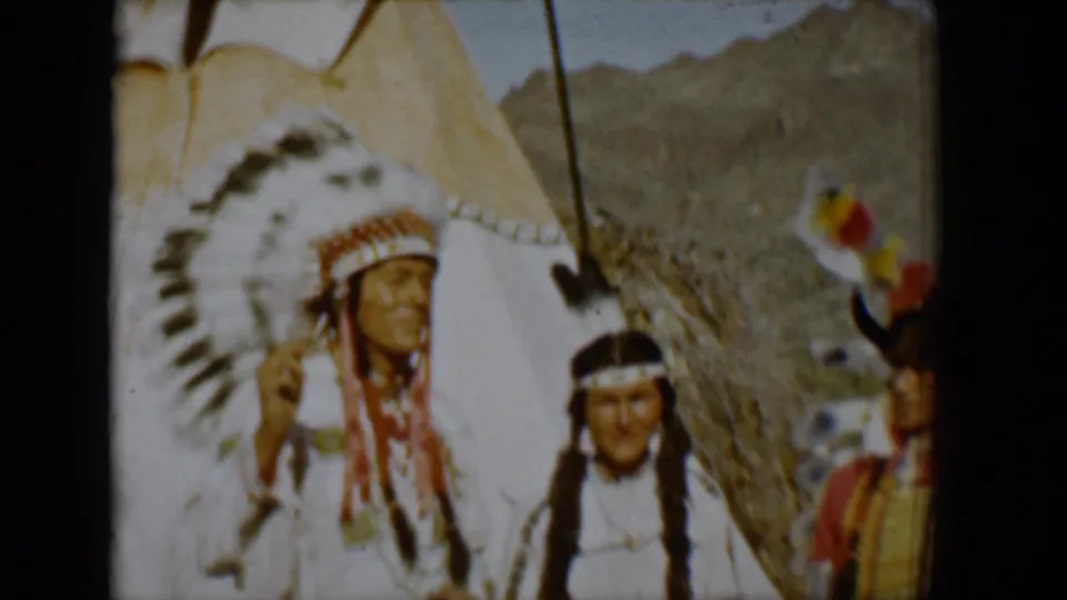 1958 Tucson home-movie frame showing adults in white regalia near a teepee, with desert terrain and a partial headdress at right.