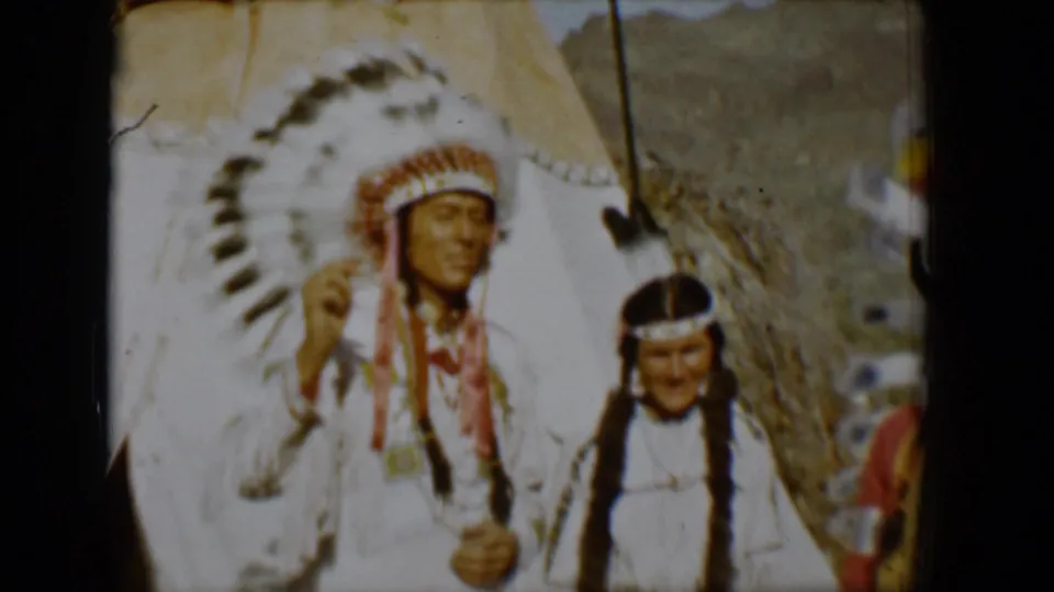 1958 Tucson frame showing a man in a large feathered headdress gesturing beside another participant in white regalia near a teepee.