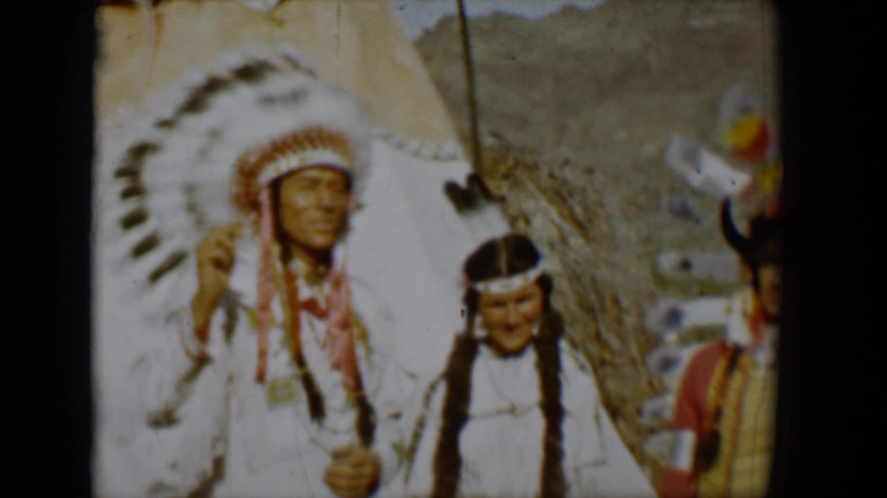 Three people in traditional regalia stand near a tepee with mountains behind them.