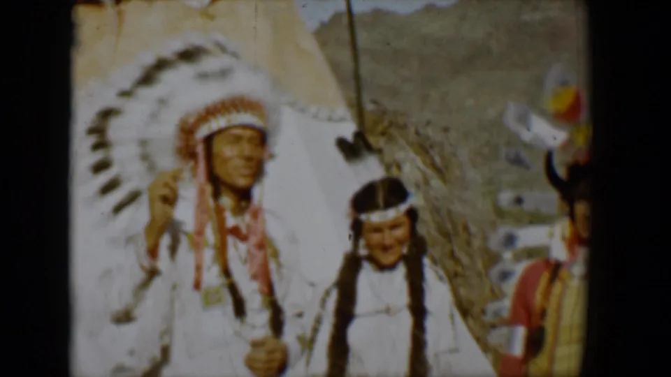 1958 Tucson home-movie frame showing a man in a feathered headdress, a woman with braids, and another figure beside a tepee.