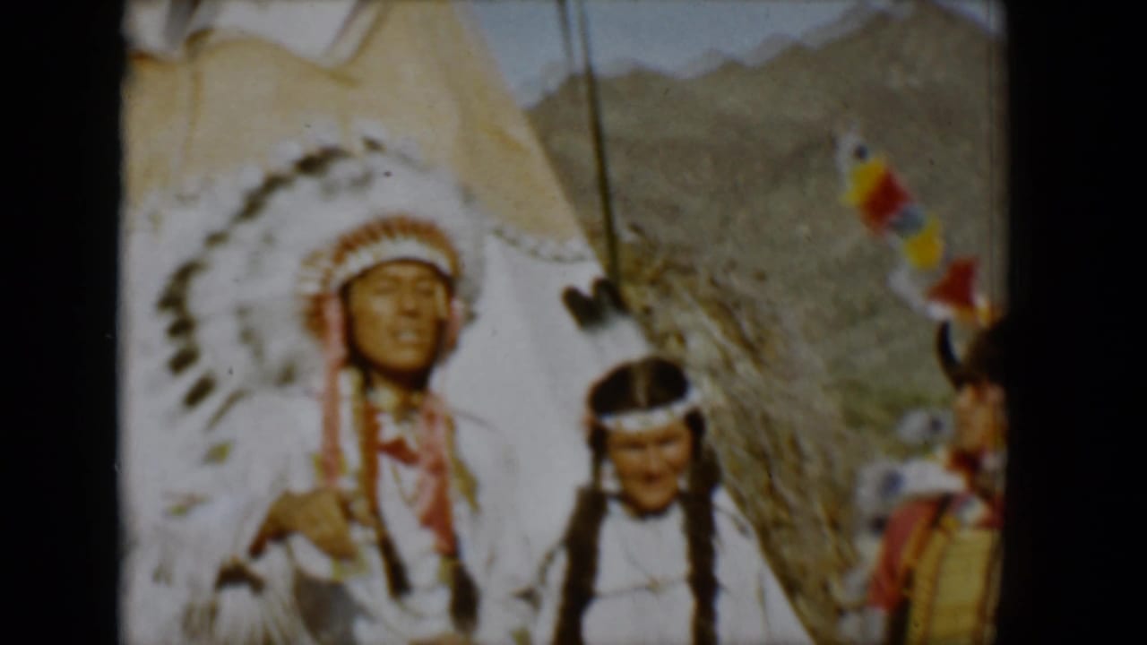 Man in a large feathered headdress stands beside a braided woman near a tepee.
