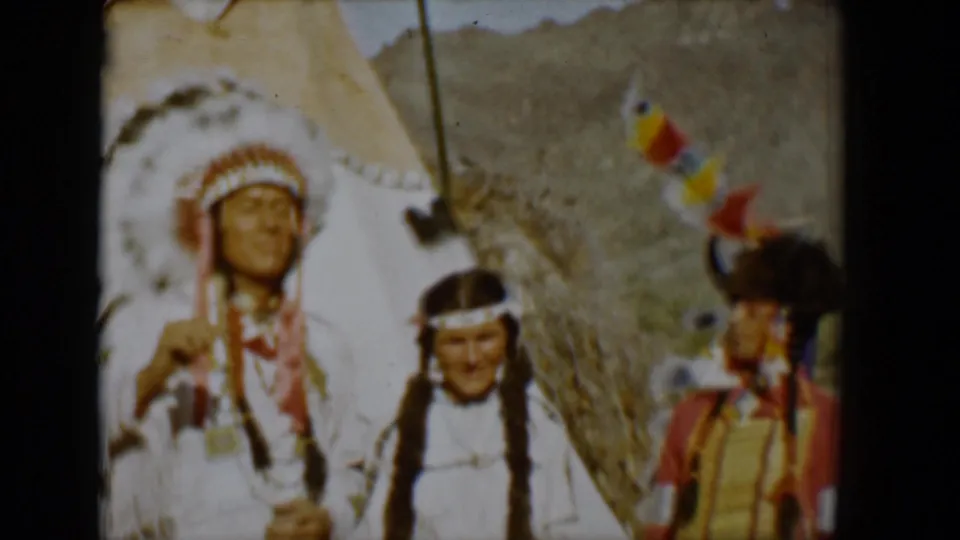 1958 Tucson home-movie frame showing regalia-clad figures near a tepee, including a man in a headdress and a figure in red and yellow.