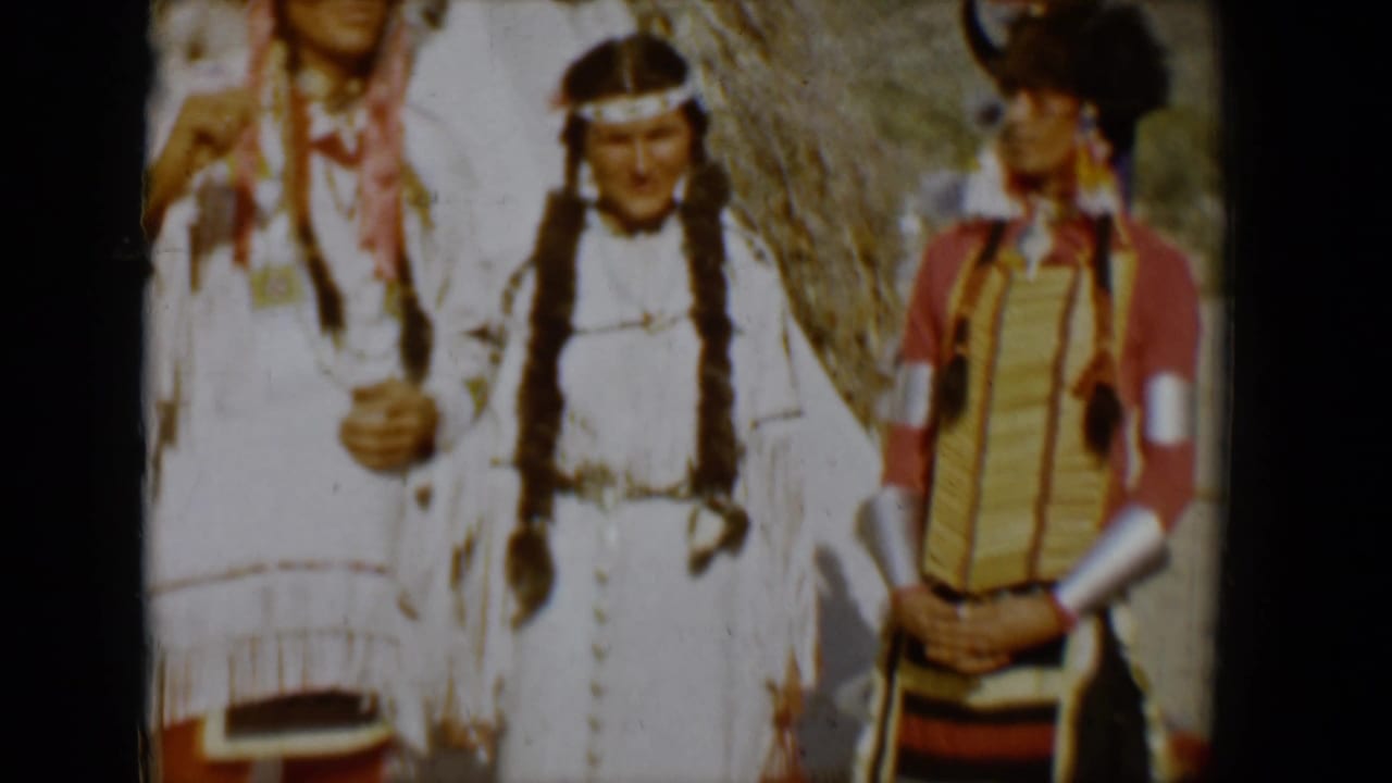 Three people standing outdoors in feathered and fringed regalia.