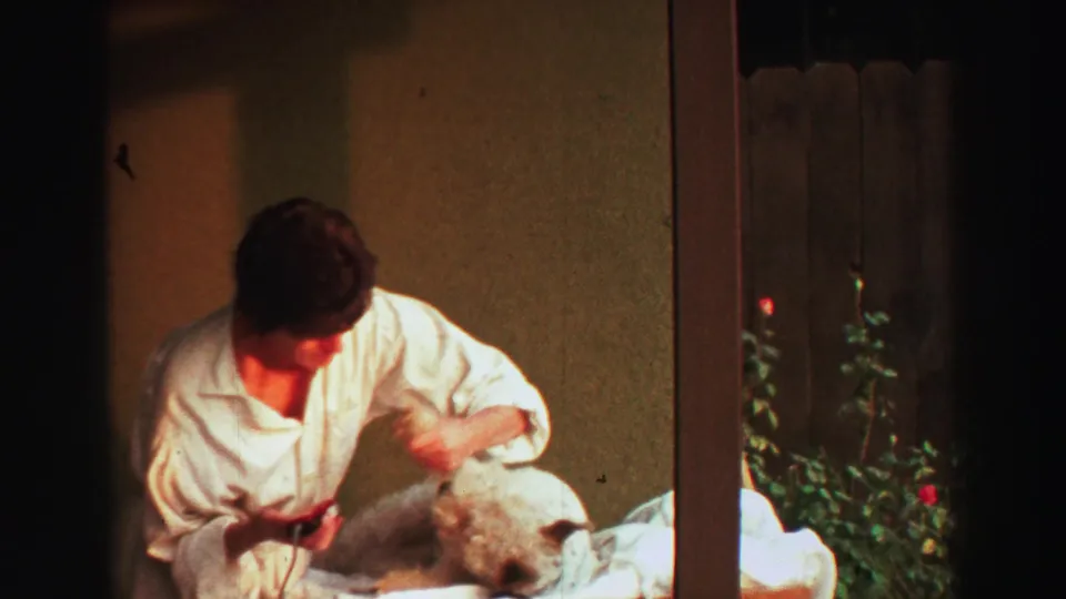 1969 Lake Tahoe home-movie frame showing a man in a white robe trimming a small fluffy dog on an outdoor table.