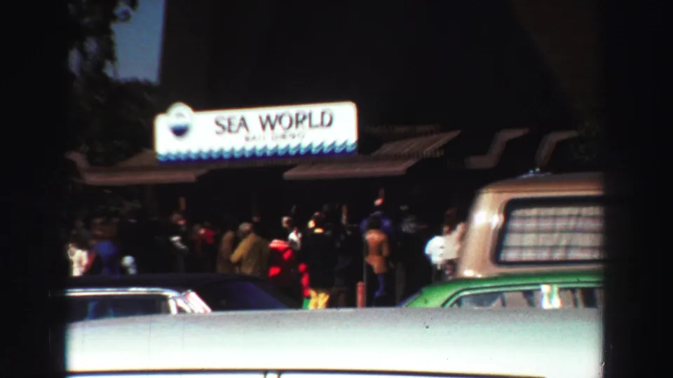 Wide 1969 visitor-shot frame of the Sea World San Diego entrance, with people gathered under the sign and vintage vehicles passing or parked nearby.