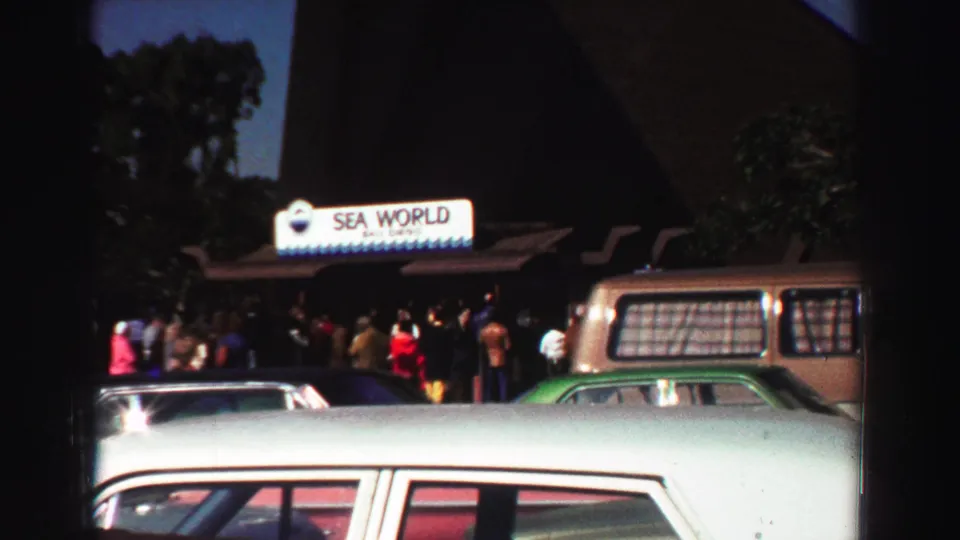 1969 8mm frame showing a Sea World entrance area with a crowd, parked cars, and a brown station wagon under a blue sky.