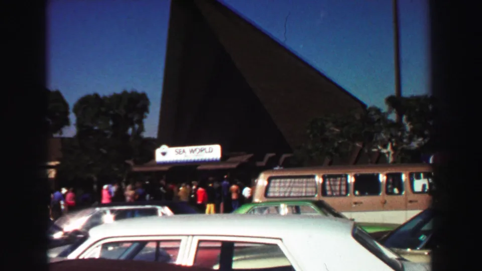 1969 8mm frame of a Sea World entrance with visitors near the sign, parked cars, a station wagon, and dark angular architecture.