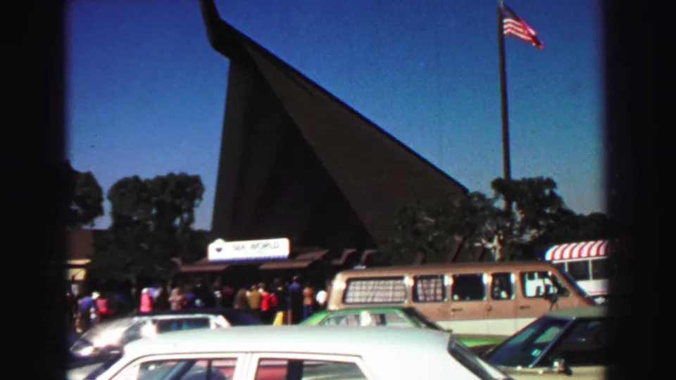 1969 8mm frame showing the Sea World entrance from the parking lot, with visitors, cars, a tall flagpole, and a steep roofline.
