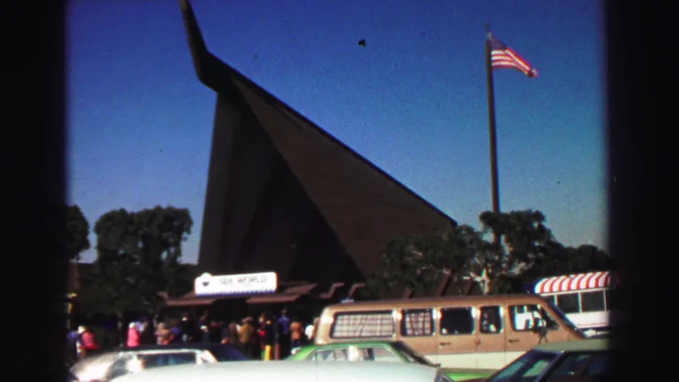 1969 8mm frame of a Sea World entrance and parking area, with visitors, period vehicles, trees, blue sky, and an American flag.