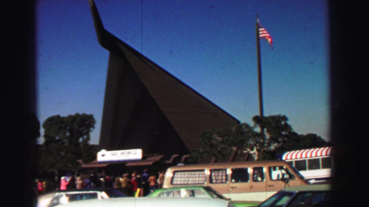 Crowd outside an A-frame building with an American flag, parked cars, and a van.