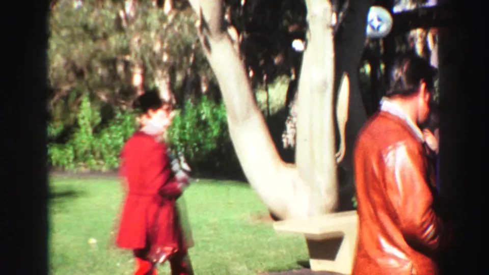 1969 Lake Tahoe home-movie frame showing adults walking in a landscaped area, with a woman in red and a man in a leather jacket.