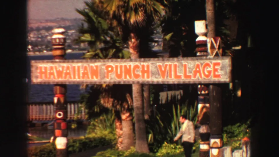1969 Lake Tahoe frame with a Hawaiian Punch Village sign, tiki-style posts, palms, a visitor, and water in the background.