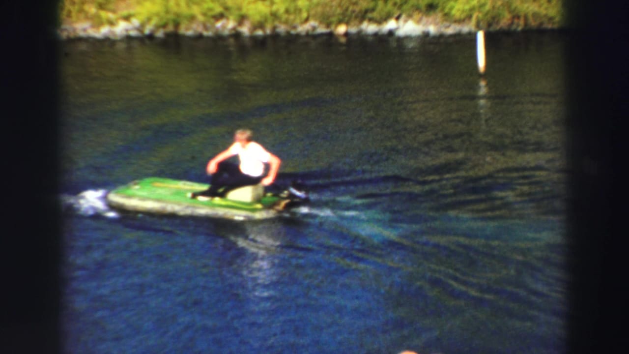 1969 Lake Tahoe home-movie frame showing a man in a white shirt on a green inflatable boat moving across dark blue water.