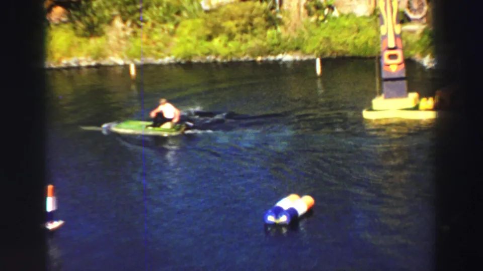 1969 Lake Tahoe frame with a man on a green inflatable boat passing colorful buoys and a floating themed structure in the distance.