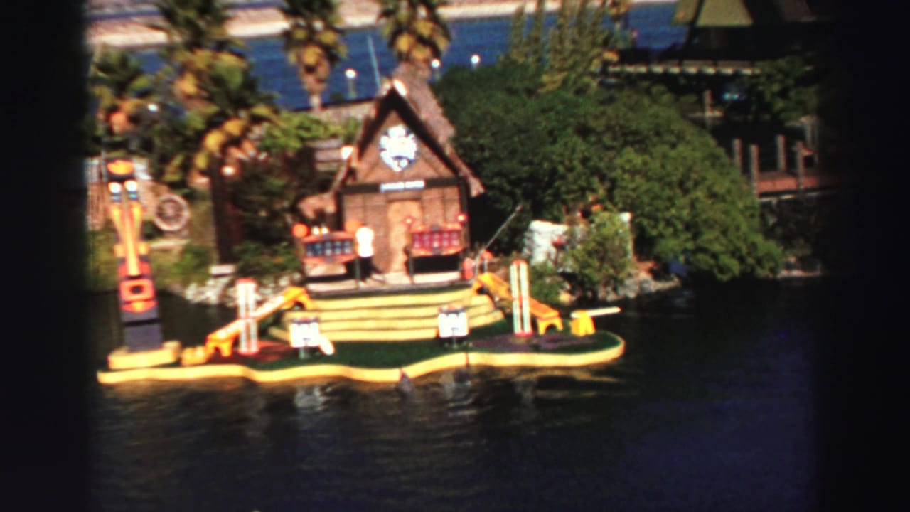 Closer view of a floating golf platform with lodge, totem-like pole, benches, greenery, and water.