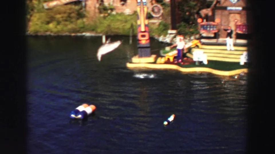 A dolphin leaps near a themed performance platform with distant people visible at the Lake Tahoe water show.