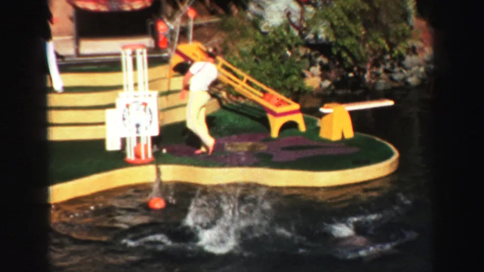 1969 Lake Tahoe frame showing a trainer on the artificial island as dolphins splash near orange balls in the pool.