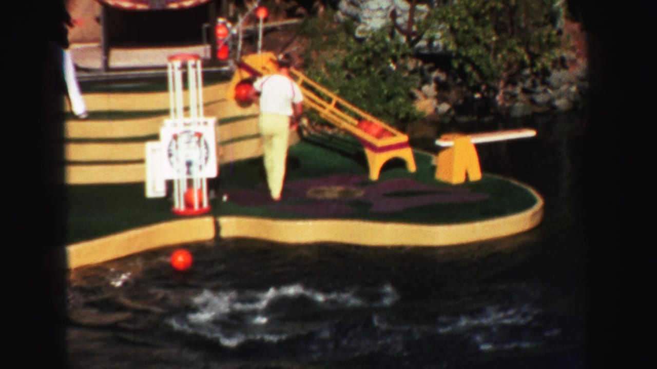 Performer stands on a floating water-show platform beside a red ball and lake water.