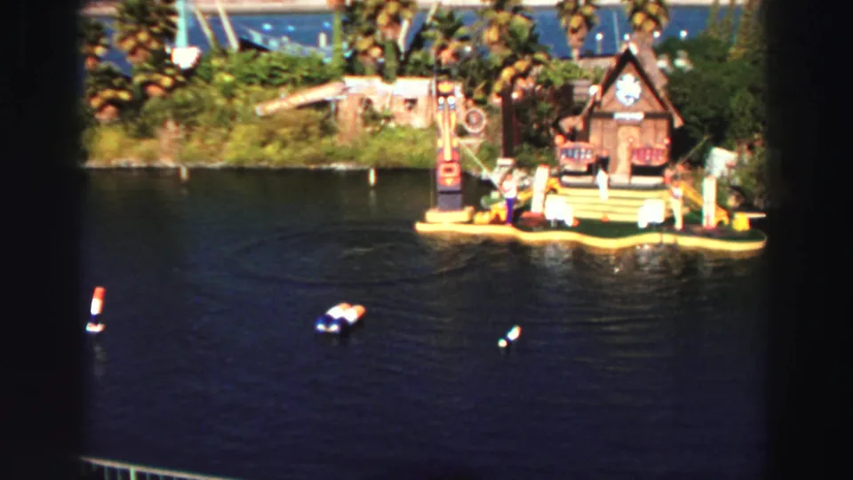 1969 Lake Tahoe frame with a colorful floating stage, cabin facade, palm trees, and people visible beside the water.