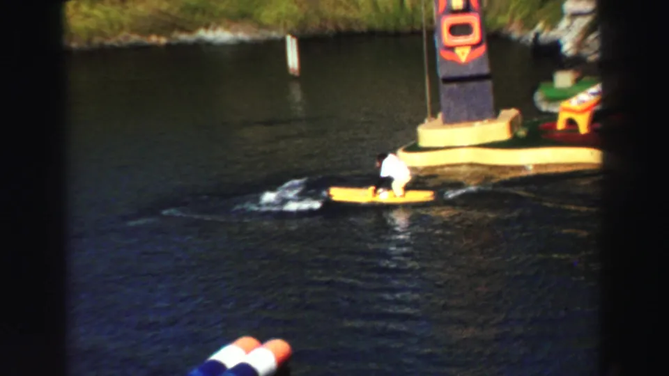 1969 Lake Tahoe home-movie frame showing a monkey in white on a yellow watercraft, with pond ripples and a colorful column behind it.