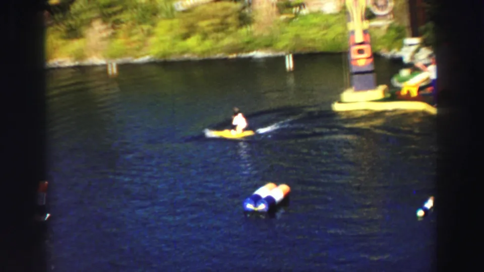 1969 Lake Tahoe frame with the monkey on a small yellow craft crossing the pond, leaving a wake near colorful floating markers.