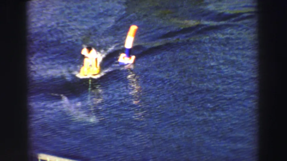 1969 Lake Tahoe frame with a rider on a yellow board circling an orange-and-white buoy while an animal shape moves below the surface.