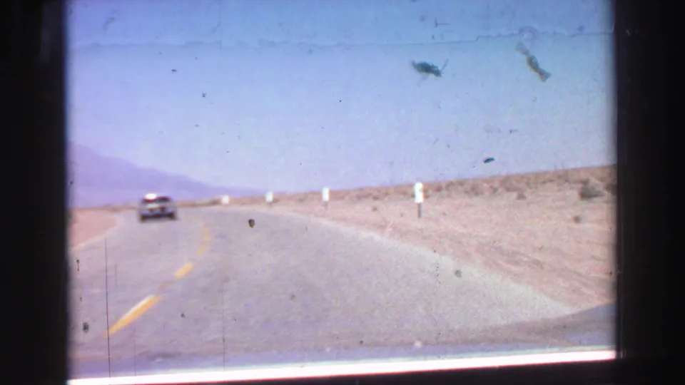 Home-movie road shot from the same 1969 travel footage, showing a car ahead on an open road with dry terrain and distant hills.