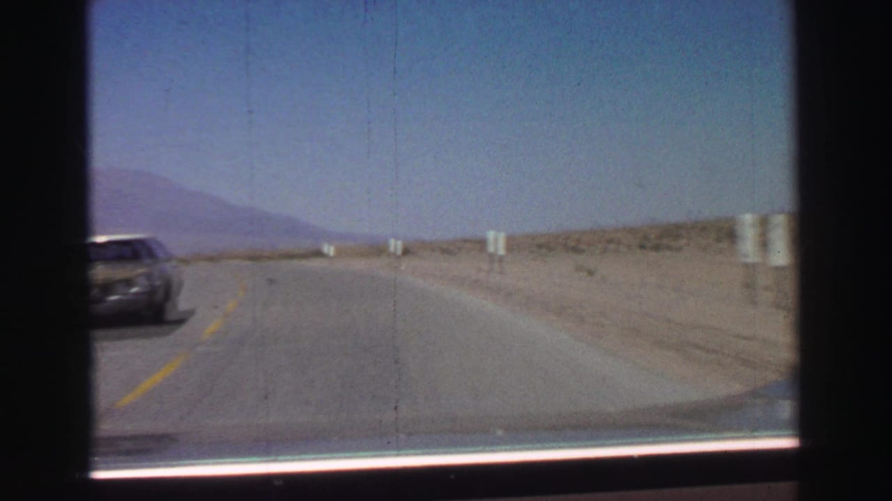 Desert highway seen through a car windshield with a passing car at left and mountains ahead.