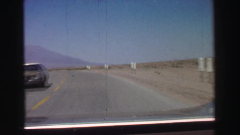 1969 8mm Lake Tahoe-area road view showing a two-lane desert highway, roadside markers, distant mountains, and an oncoming car passing left.