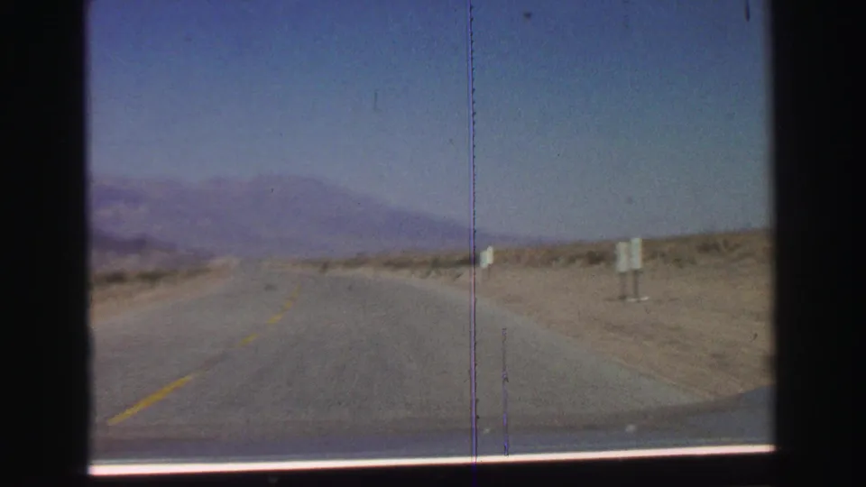 1969 8mm road-trip frame showing an open two-lane highway through arid terrain with white roadside markers and mountains ahead.