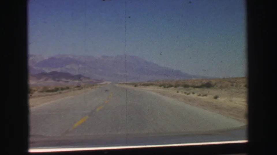 1969 8mm Lake Tahoe-area driving view with a straight two-lane road, low desert vegetation, and hazy mountains in the distance.