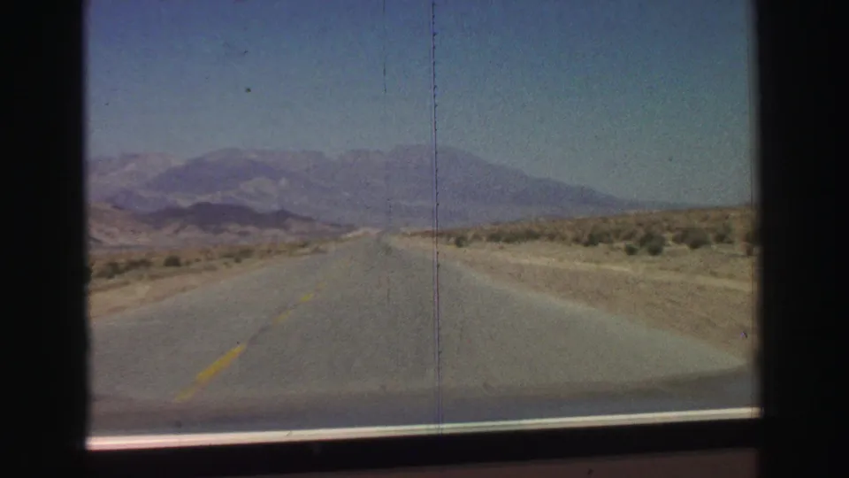 1969 8mm driving view near Lake Tahoe showing an empty highway, dry roadside land, white markers, and mountains under a clear sky.