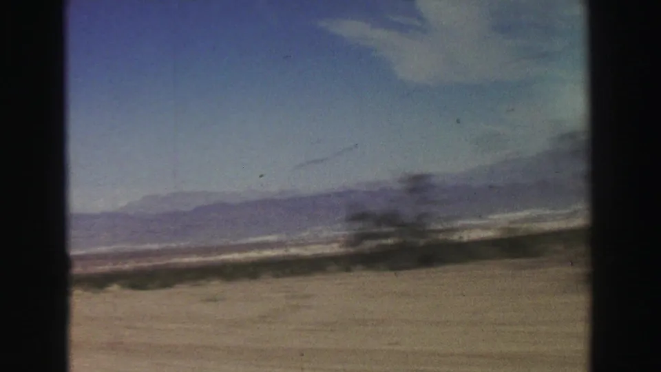 A moving-car view captures arid roadside brush, open desert floor, and distant California mountain ridges in soft 8mm color.