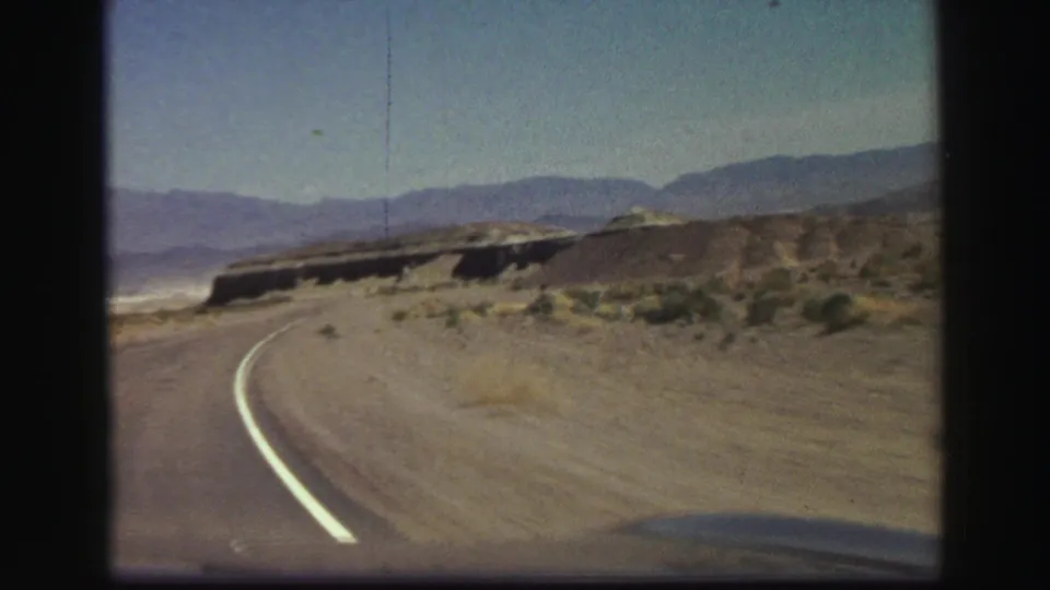 A curving road and painted lane line lead through an arid California landscape toward hills and distant mountains in 1969 8mm footage.