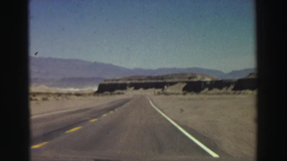Vintage 8mm view from a moving vehicle on a two-lane highway, with sandy roadside and mountains in the background.