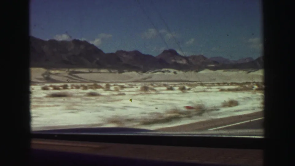A moving-car view across bright desert flats toward dark mountains, with dashboard and window framing visible in the 8mm frame.