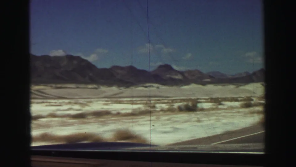 Vintage 8mm view from a car across a sunlit desert basin with sparse brush, road edge, and mountains in the distance.