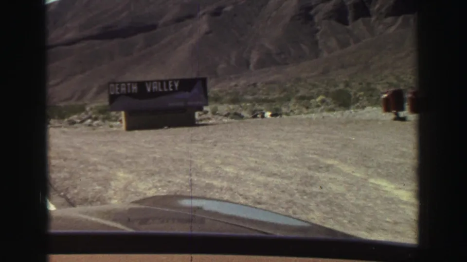 Approaching view of a black Death Valley sign near a rocky hillside, filmed from inside a car on 8mm footage.