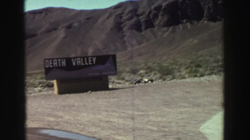 The Death Valley sign remains visible from a car-window perspective, with barren roadside ground and dark hills behind it.