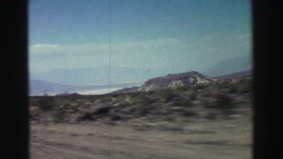 A moving-car 8mm view shows desert plants, open road edge, low hills, and pale distant mountains after passing the sign.