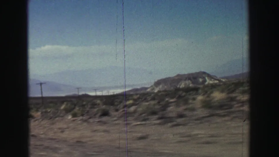 A later frame shows roadside desert vegetation, utility poles, pale mountains, and a rounded hill filmed from the moving car.