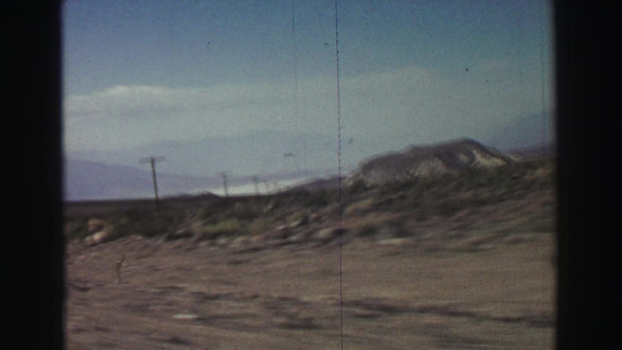 Desert roadside with utility poles, low shrubs, and distant rocky mountains under blue sky.
