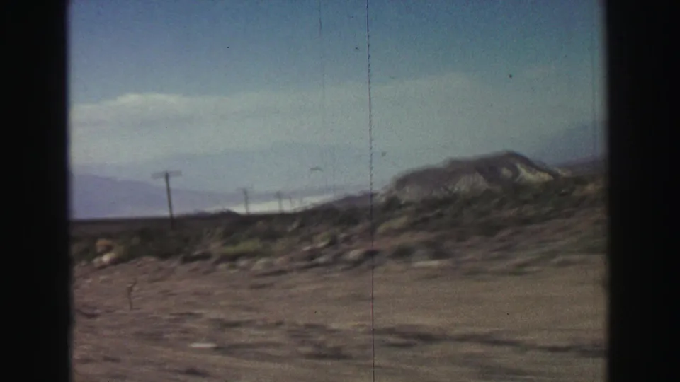 1969 8mm Lake Tahoe-area road-trip view of arid roadside terrain, utility poles, scrub, and distant mountains seen from a moving vehicle.