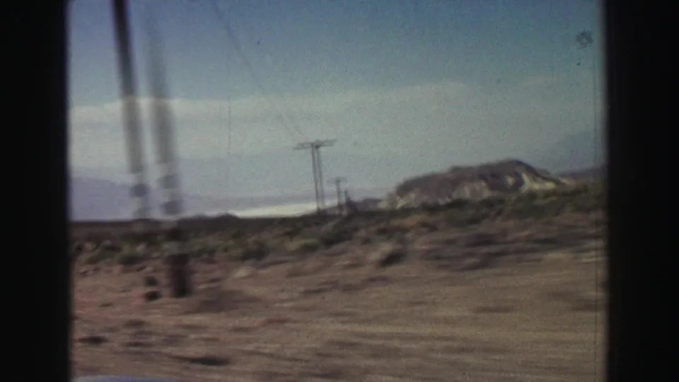 Moving-car view in 1969 8mm footage showing dry roadside scrub, utility poles, and a low rocky mountain near Lake Tahoe, California.