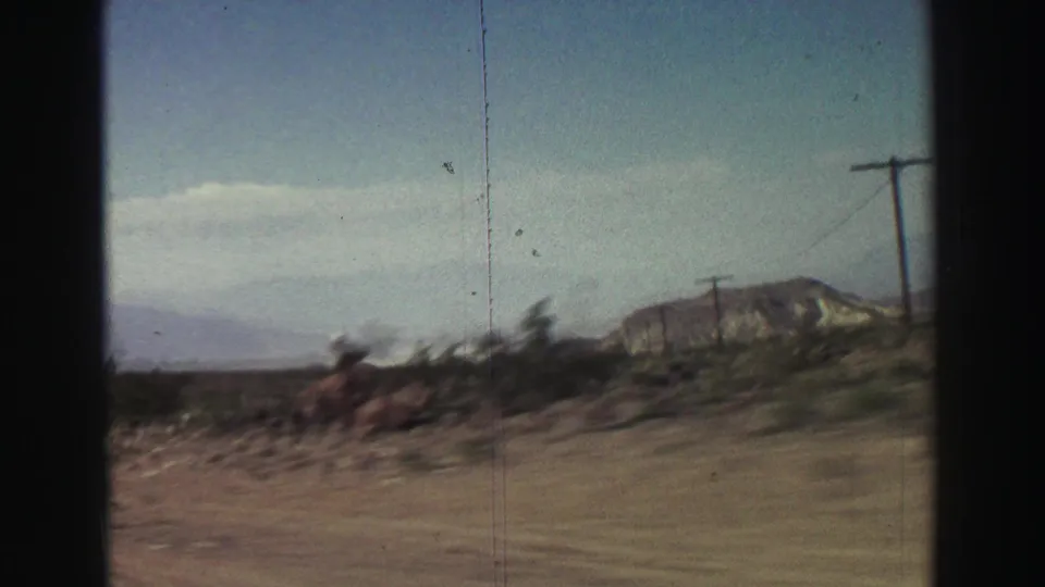 Passenger-side 8mm view of sparse desert vegetation, roadside poles, and hazy mountain ridges during a 1969 California road trip.