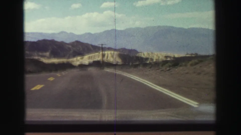 Forward-facing 1969 8mm road view near Lake Tahoe, with a two-lane highway, roadside poles, pale hills, and distant mountains.