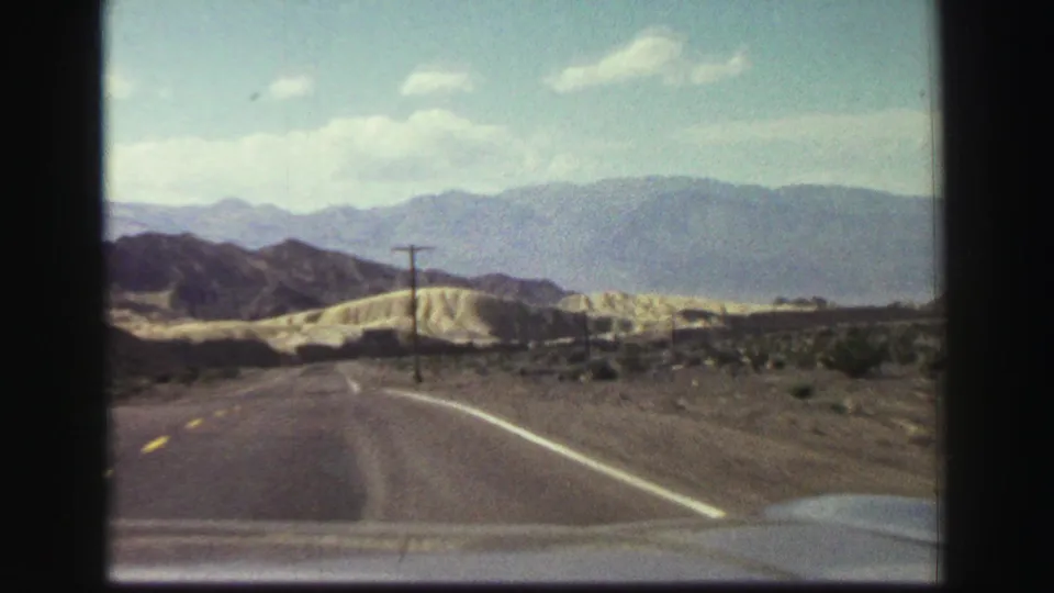 1969 8mm windshield view near Lake Tahoe showing a paved road, yellow center line, white edge line, dry scrub, and mountains beyond.