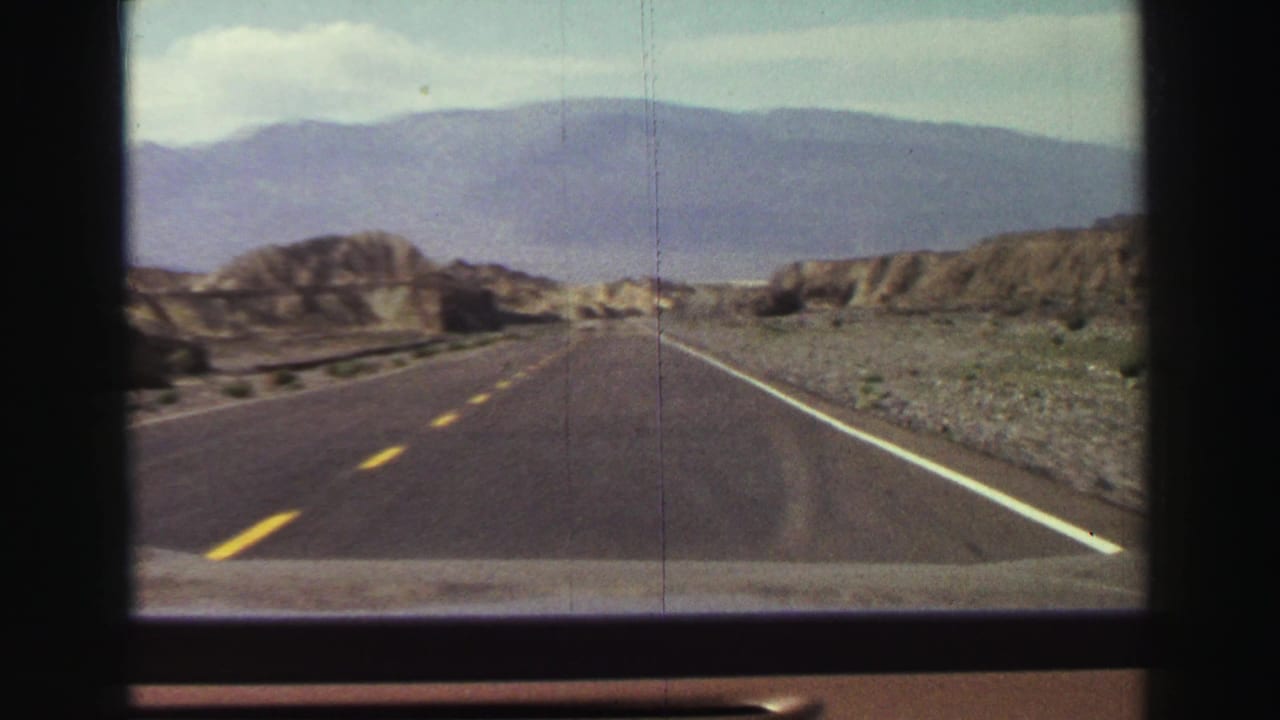 Two-lane highway stretching toward rocky hills and distant mountains under a pale blue sky.